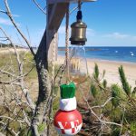 applique murale lumineuse. Composé d'une potence en bois flotté. D'une douille laiton, d'une ampoule allongée à filament. De 4 flotteurs de pêche de couleur vert, blanc et rouge suspendu à une corde en jute. L'applique est accrochée à un arbre devant une plage