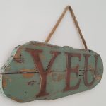 cadre mural en bois peint en vert patiné. Grandes lettres en bois effet métal rouillé peintes en rouille. Accrochée sur mur blanc avec grosse corde en jute.