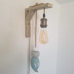 applique murale lumineuse. Composé d'une potence en bois flotté. D'une douille laiton, d'une ampoule allongée à filament. De 3 flotteurs de pêche dans les tons bleu ciel, gris et blanc, suspendus à une corde en jute. On peut lire YE sur un flotteur. Applique accrochée le long d'un mur blanc