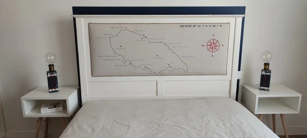tête de lit en bois blanc capitonnée. La carte de l'île d'Yeu avec le nom des plages a été dessiné sur le tissu. On voit une rose des vents en rouge. 2 chevets blancs de chaque côté du lit et 2 lampes en bois bleu marine posées dessus