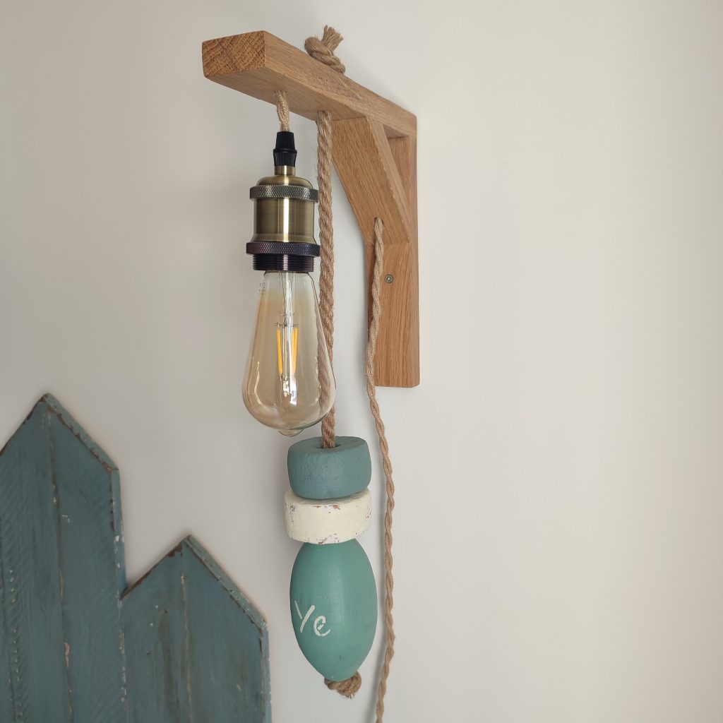 applique murale lumineuse. Composé d'une potence en bois chêne naturel. D'une douille laiton, d'une ampoule allongée à filament. De 3 flotteurs de pêche dans les tons vert céladon et blanc, suspendus à une corde en jute. On peut lire YE sur un flotteur. Applique accrochée le long d'un mur blanc dans une chambre avec tête de lit et chevet bleu.
