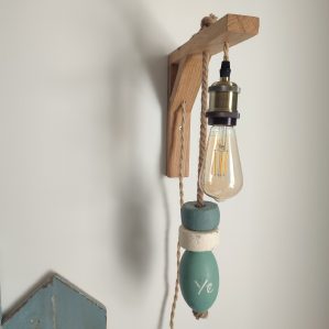 applique murale lumineuse. Composé d'une potence en bois chêne naturel. D'une douille laiton, d'une ampoule allongée à filament. De 3 flotteurs de pêche dans les tons vert céladon et blanc, suspendus à une corde en jute. On peut lire YE sur un flotteur. Applique accrochée le long d'un mur blanc dans une chambre avec tête de lit et chevet bleu.