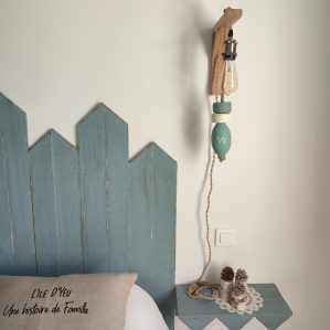 applique murale lumineuse. Composé d'une potence en bois chêne naturel. D'une douille laiton, d'une ampoule allongée à filament. De 3 flotteurs de pêche dans les tons vert céladon et blanc, suspendus à une corde en jute. On peut lire YE sur un flotteur. Applique accrochée le long d'un mur blanc dans une chambre avec tête de lit et chevet bleu.