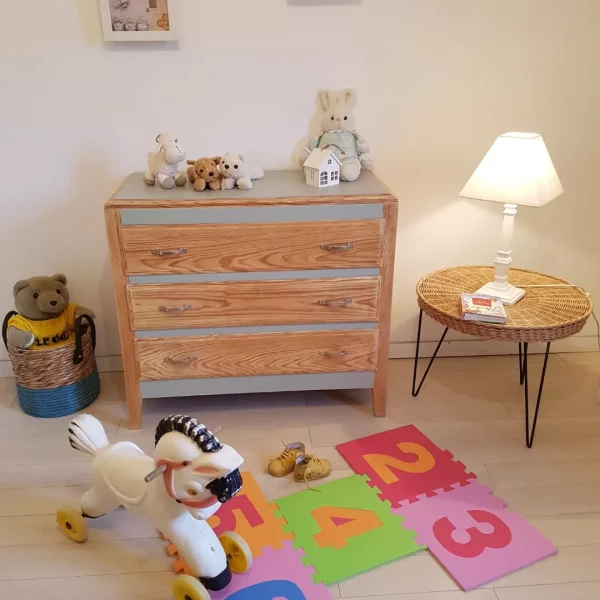 commode 3 tiroirs en pin peinte partiellement en vert sauge . Présentée dans une chambre de bébé. Jouets, peluches, tapis au sol