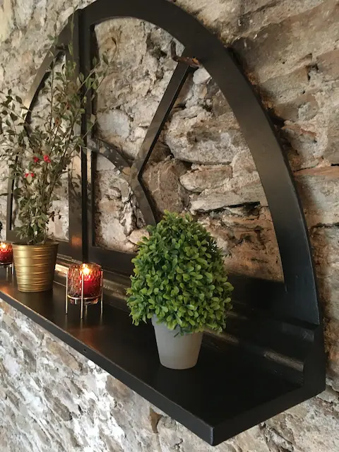 détournement d'un chassis de fenêtre cintré en bois noir en étagère murale. La fenêtre est fixée sur un mur en pierres apparentes. De la décoration de Noël est posé sur l'étagère. Ambiance très chaleureuse.