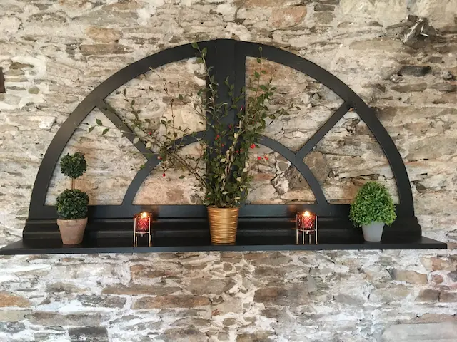 détournement d'un chassis de fenêtre cintré en bois noir en étagère murale. La fenêtre est fixée sur un mur en pierres apparentes. De la décoration de Noël est posé sur l'étagère. Ambiance très chaleureuse.