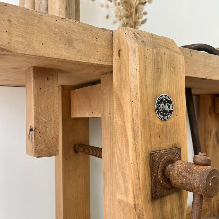 Partie d'un établi ancien en chêne massif. On voit la presse. Un bouquet de fleurs séchées est posé sur l'établi