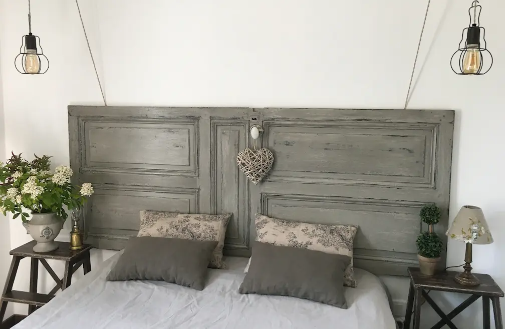 porte ancienne rénovée. Peinte en kaki, patinée pour donner du charme. Détournée en tête de lit. On voit la chambre, le lit. Les draps sont blancs, les coussins sont beige et kaki. 2 tables de chevets ornent la tête de lit. Des ampoules en suspensions sont accrochées au plafond et permettent d'éclairer la chambre