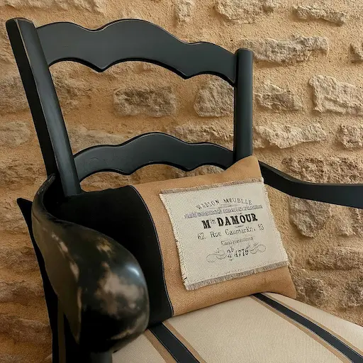 fauteuil de campagne peint en noir. Un coussin en tissu noir et jute est posé sur l'assise du fauteuil . Fauteuil devant un mur en pierres apparentes