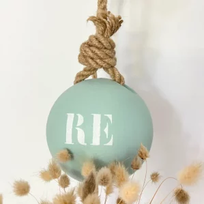on voit un flotteur de pêche rond peint en bleu. Il est suspendu à une corde en jute et il est inscrit dessus RE. Au premier plan il y a des fleurs des dunes.