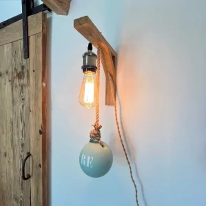 applique murale lumineuse accrochée sur mur blanc. Elle est composée d'une potence en bois clair, douille laiton, ampoule à filament, flotteur de pêche peint en bleu ciel sur lequel est inscrit RE. On voit une vieille porte en bois sur rail noir à côté de l'applique.