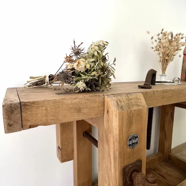 Partie d'un établi ancien en chêne massif. On voit la presse. Un bouquet de fleurs séchées est posé sur l'établi