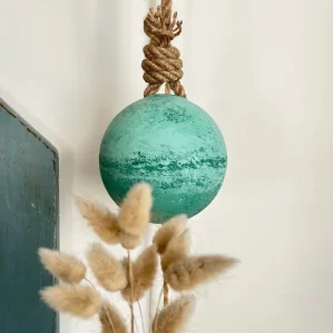 on voit un flotteur de pêche rond peint dans différents tons de vert. Il est suspendu à une corde en jute. Au premier plan il y a des fleurs des dunes.
