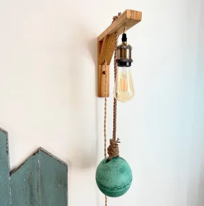 applique murale lumineuse accrochée sur mur blanc. Elle est composée d'une potence en bois clair, douille laiton, ampoule à filament, flotteur de pêche peint en vert.