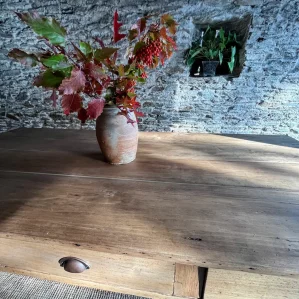 plateau de table de ferme. Présentée devant un mur en pierres apparentes. Un bouquet végétal automnal est disposée dans un pot en terre au centre de la table.