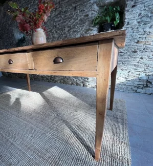 gros plan d'un tiroir fermé d'une table de ferme présentée devant un mur en pierres apparentes. Un bouquet végétal automnal est disposée dans un pot en terre au centre de la table.
