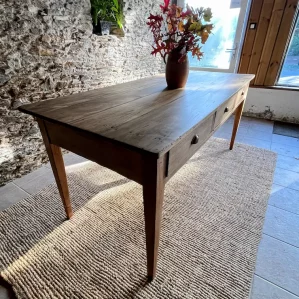 table de ferme en bois massif, 3 tiroirs, présentée devant un mur en pierres apparentes. Un bouquet végétal automnal est disposée dans un pot en terre au centre de la table.