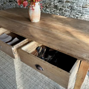 table de ferme en bois massif présentée devant un mur en pierres apparentes. Un bouquet végétal automnal est disposée dans un pot en terre au centre de la table.. 2 tiroirs sont ouverts.