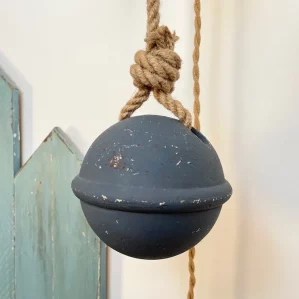 on voit au premier plan un flotteur de pêche rond peint dans en gris bleuté. Il est suspendu à une corde en jute. Au second plan on voit des planches en bois fixées au mur et peintes en vert céladon