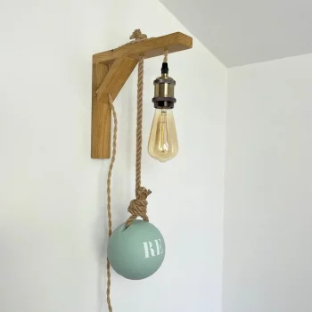 applique murale lumineuse accrochée sur mur blanc. Elle est composée d'une potence en bois clair, douille laiton, ampoule à filament, flotteur de pêche peint en bleu ciel sur lequel est inscrit RE. On voit une vieille porte en bois sur rail noir à côté de l'applique.